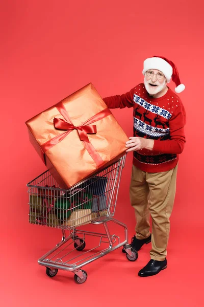 Homme âgé souriant en pull et chapeau santa tenant présent près du panier sur fond rouge — Photo de stock