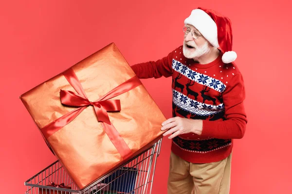 Positive senior man in santa hat and sweater holding gift with bow near shopping cart isolated on red — Stock Photo