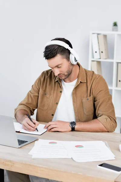 Businessman in headphones writing in notebook while sitting at workplace with digital devices — Stock Photo