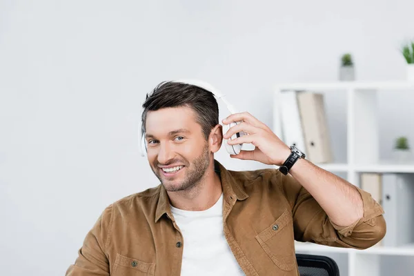 Homme d'affaires joyeux décoller écouteurs tout en regardant la caméra avec un bureau flou sur fond — Photo de stock