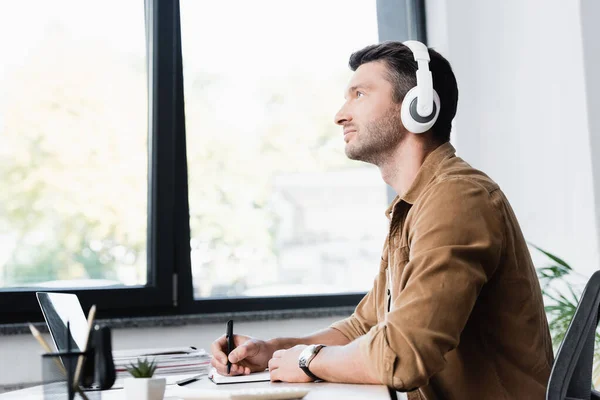 Vista lateral del hombre de negocios en auriculares con lápiz mirando hacia otro lado mientras está sentado en el lugar de trabajo con ventana borrosa en el fondo - foto de stock