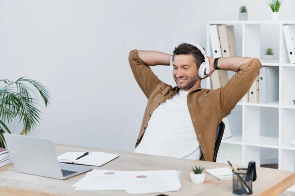 Hombre de negocios alegre con las manos detrás de la cabeza mirando a la computadora portátil mientras está sentado en el lugar de trabajo en la oficina - foto de stock