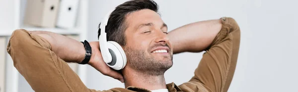 Homme d'affaires souriant dans un casque, les mains derrière la tête et écoutant de la musique sur fond flou, bannière — Photo de stock