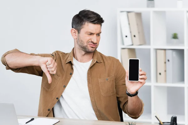 Dissatisfied businessman with thumb down, holding smartphone with blank screen at workplace on blurred background — Stock Photo