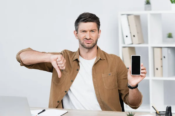 Uomo d'affari insoddisfatto con il pollice verso il basso, guardando la fotocamera mentre tiene lo smartphone sul posto di lavoro su sfondo sfocato — Foto stock