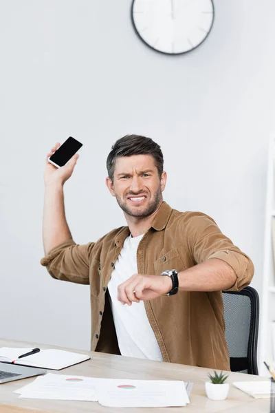 Homem de negócios irritado olhando para a câmera enquanto se livra do smartphone no local de trabalho — Fotografia de Stock