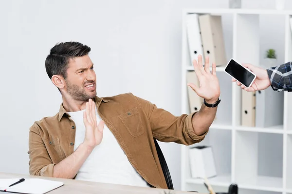 Hombre de negocios irritado con gesto de stop mirando el teléfono inteligente en la mano del hombre cerca del lugar de trabajo - foto de stock