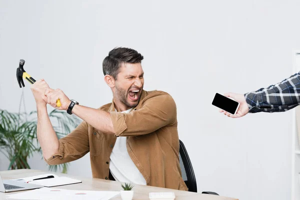Aggressive businessman with hammer shouting while sitting near smartphone in hand of man near workplace — Stock Photo