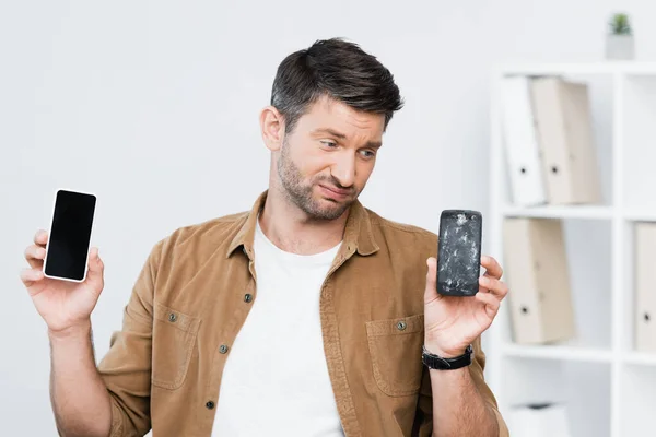 Hombre de negocios insatisfecho con teléfono inteligente mirando teléfono celular roto sobre fondo borroso - foto de stock