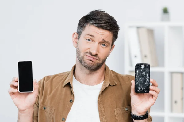 Незадоволений бізнесмен дивиться на камеру, тримаючи розбитий смартфон і мобільний телефон з порожнім екраном — стокове фото