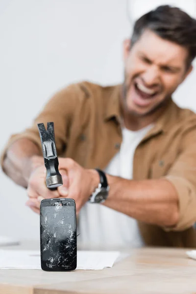 Hombre de negocios agresivo gritando mientras sostiene el martillo cerca de teléfono inteligente aplastado en la mesa sobre fondo borroso - foto de stock