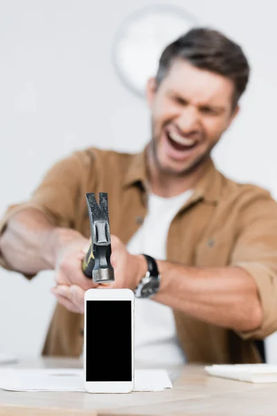Hombre de negocios agresivo gritando mientras sostiene el martillo cerca del teléfono inteligente con pantalla en blanco en la mesa sobre un fondo borroso - foto de stock