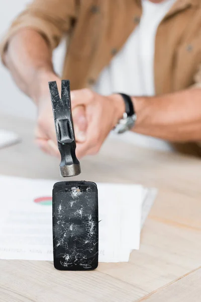 Vue recadrée d'un homme d'affaires tenant un marteau près d'un smartphone fracassé sur une table sur fond flou — Photo de stock