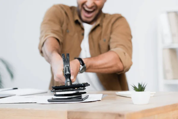 Vista ritagliata di gridare uomo d'affari cracking mucchio di smartphone con martello sul tavolo su sfondo sfocato — Foto stock