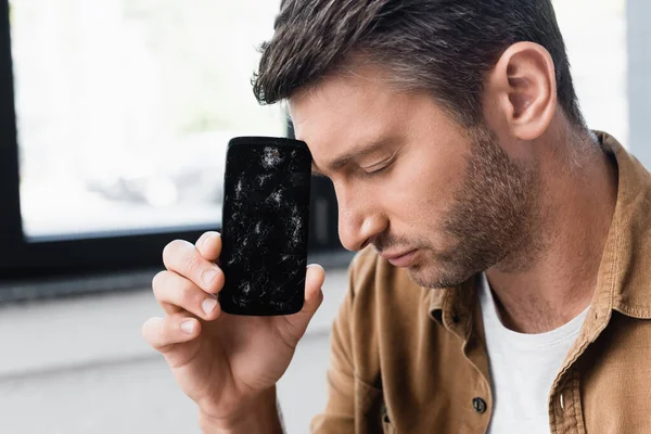 Vista laterale di uomo d'affari sconvolto con gli occhi chiusi in possesso di smartphone fracassato vicino alla fronte su sfondo sfocato — Foto stock