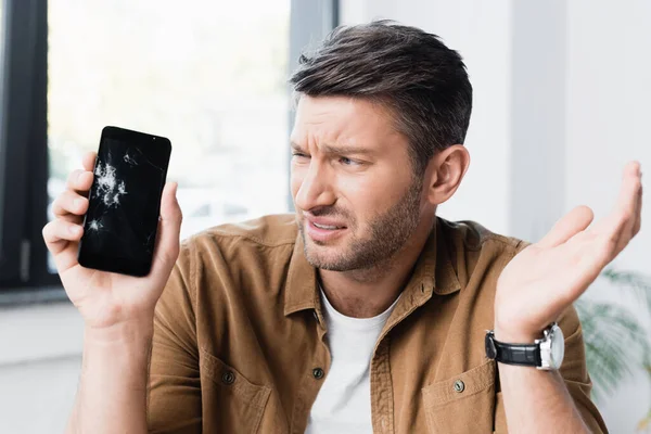 Worried businessman gesturing while looking at smashed smartphone on blurred background — Stock Photo