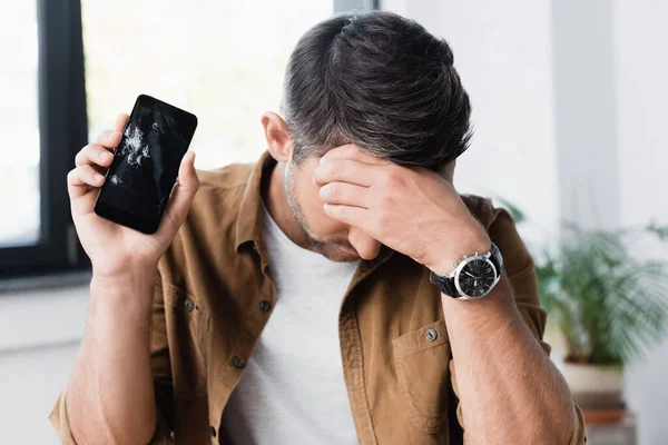 Disappointed businessman with hand on forehead, holding smashed smartphone on blurred background — Stock Photo