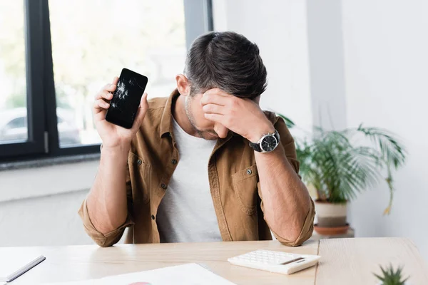 Empresário decepcionado com smartphone esmagado sentado no local de trabalho em fundo embaçado — Fotografia de Stock