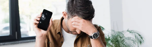 Sad businessman with hand on forehead, holding smashed smartphone on blurred background, banner — Stock Photo