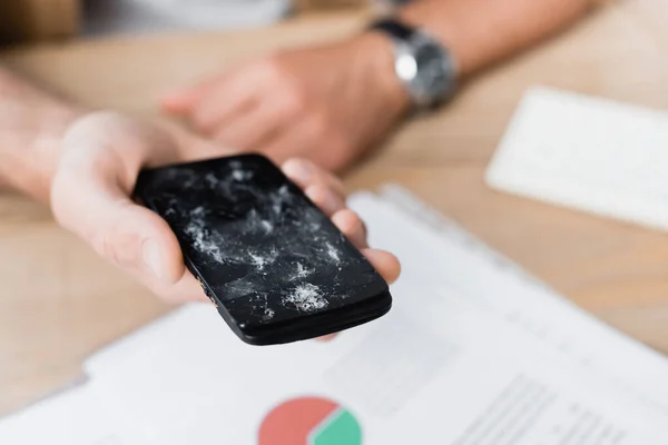 Cropped view of businessman holding smashed smartphone with blurred workplace on background — Stock Photo