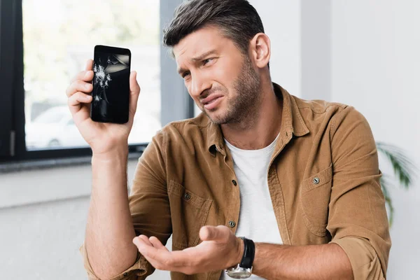 Upset businessman gesturing while looking at smashed smartphone with blurred window on background — Stock Photo