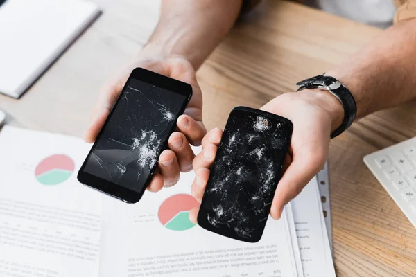 Vue recadrée d'un homme d'affaires tenant un smartphone brisé près du lieu de travail sur fond flou — Photo de stock