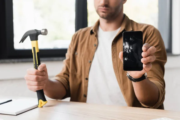 Cropped view of businessman with hammer showing damaged smartphone while sitting at workplace on blurred background — Stock Photo