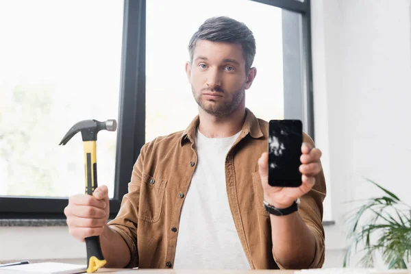 Trauriger Geschäftsmann mit Hammer blickt in Kamera und zeigt beschädigtes Smartphone mit verschwommenem Fenster im Hintergrund — Stockfoto