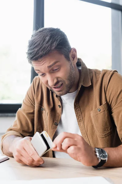 Dissatisfied businessman disassembling smashed smartphone while sitting at workplace on blurred background — Stock Photo