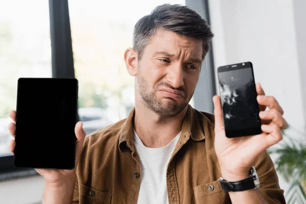 Triste hombre de negocios sosteniendo el teléfono inteligente con pantalla en blanco mientras mira el teléfono celular roto en un fondo borroso - foto de stock