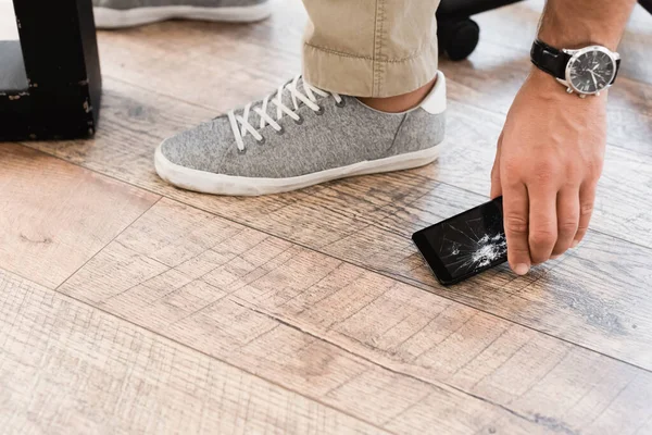 Ausgeschnittene Ansicht eines Geschäftsmannes, der beschädigtes Smartphone vom Holzboden holt — Stockfoto