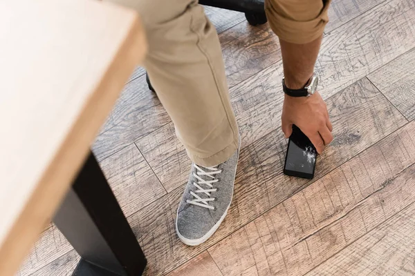 Vista ritagliata di uomo d'affari che prende cellulare fracassato dal pavimento in legno con tavolo sfocato in primo piano — Foto stock