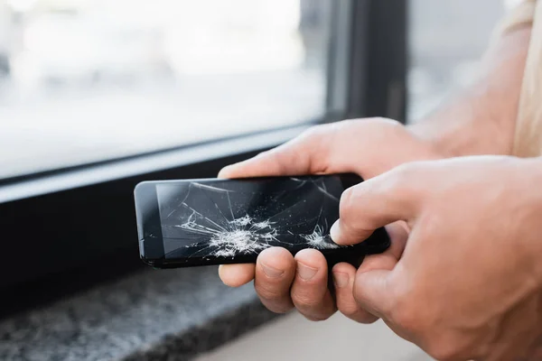 Cropped view of businessman touching smashed touchscreen of smartphone with big finger on blurred background — Stock Photo