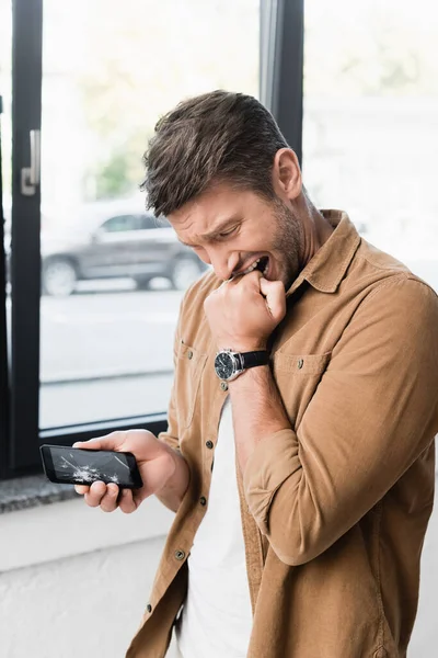 Frightened businessman biting fist while looking at smashed smartphone with blurred window on background — Stock Photo