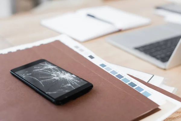 Nahaufnahme eines zertrümmerten Smartphones auf einem Papierstapel mit verschwommenem Arbeitsplatz im Hintergrund — Stockfoto