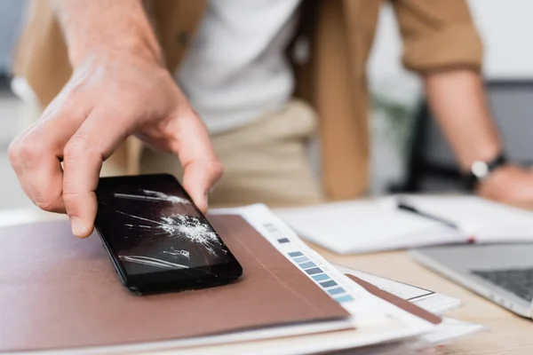 Cropped view of businessman taking smartphone with crack from pile of paperwork on blurred background — Stock Photo