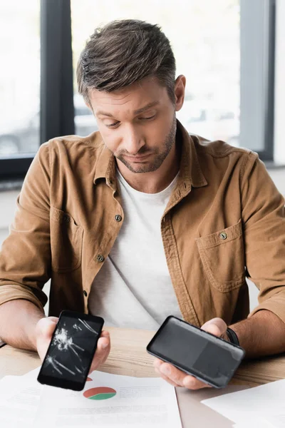 Скептик-бизнесмен смотрит на разбитые разобранные смартфоны, сидя за столом на размытом фоне — стоковое фото
