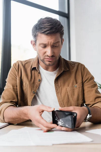 Грустный бизнесмен разбирает поврежденный смартфон, сидя на рабочем месте на размытом фоне — стоковое фото