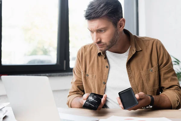 Serious businessman with disassembled smashed smartphone, looking at laptop at workplace on blurred background — Stock Photo