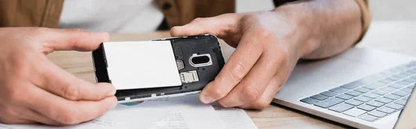 Vista recortada del hombre de negocios poniendo la batería en el teléfono móvil roto en la mesa, bandera - foto de stock