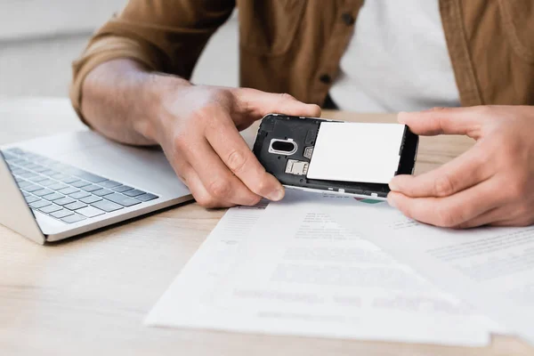 Cropped view of businessman putting battery in mobile phone at workplace with paperwork on blurred foreground — Stock Photo