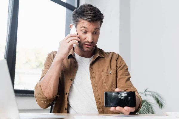 Uomo d'affari con smartphone fracassato che parla sul cellulare mentre è seduto sul posto di lavoro su sfondo sfocato — Foto stock