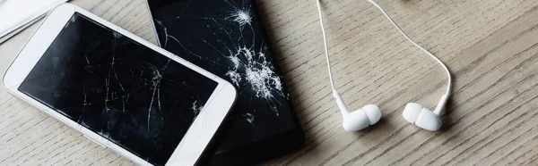 Top view of earphones with smashed smartphone on wooden background, banner — Stock Photo