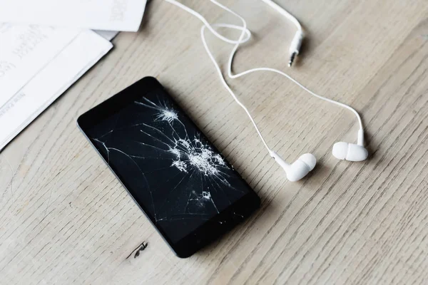 Vista de cerca del teléfono celular roto con auriculares y hojas de papel sobre fondo de madera - foto de stock