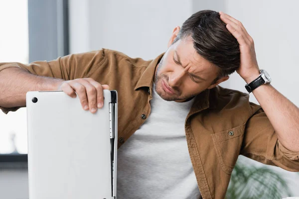 Worried businessman thinking while looking at broken laptop on blurred background — Stock Photo