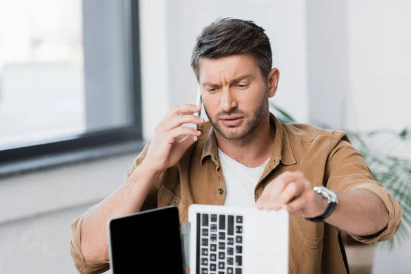 Hombre de negocios escéptico mirando a la computadora portátil rota mientras habla en el teléfono móvil en un fondo borroso - foto de stock