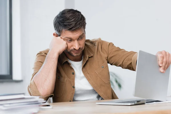 Disappointed businessman thinking while looking at damaged laptop at workplace on blurred foreground — Stock Photo
