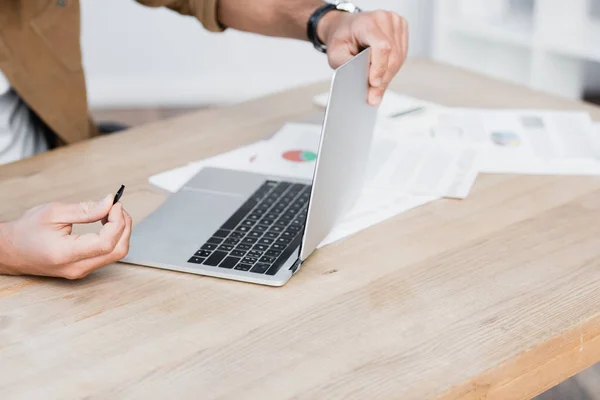 Ausgeschnittene Ansicht eines Geschäftsmannes mit Detail, der einen kaputten Laptop mit Papieren auf verschwommenem Hintergrund am Tisch hält — Stockfoto