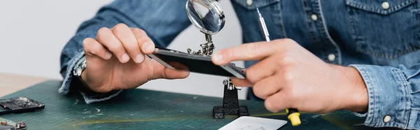 Vue recadrée du réparateur tenant une partie démontée du téléphone mobile près de la loupe sur le lieu de travail, bannière — Photo de stock