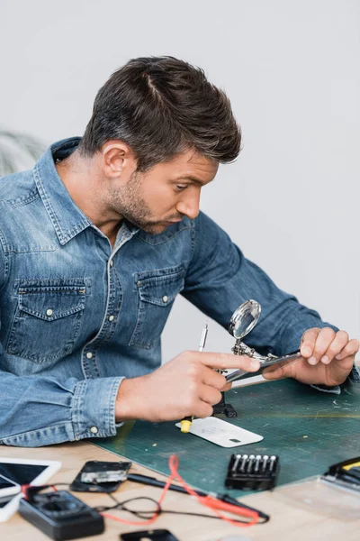 Reparaturarbeiter mit Schraubenzieher blickt durch Lupe auf zerlegtes Teil des Mobiltelefons am Tisch auf verschwommenem Vordergrund — Stockfoto